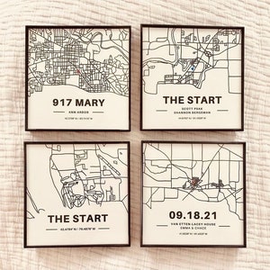 May include: Four black framed prints with white backgrounds and black outlines of maps. The top left print has the text "917 MARY" and "ANN ARBOR" with coordinates "42.2700&deg;N / 83.7415&deg;W". The top right print has the text "THE START" and "SCOTT PSAK SHANNON BERGEMAN" with coordinates "44.8763&deg;N / 91.4330&deg;W". The bottom left print has the text "THE START" and coordinates "42.4784&deg;N / -76.4878&deg;W". The bottom right print has the text "09.18.21" and "VAN ETTEN-LACEY HOUSE EMMA & CHACE" with coordinates "41.9226&deg;N / -91.4222&deg;W".