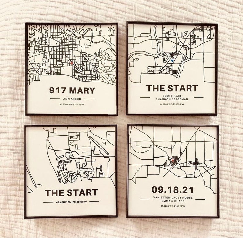 May include: Four black framed prints with white backgrounds and black outlines of maps. The top left print has the text "917 MARY" and "ANN ARBOR" with coordinates "42.2700&deg;N / 83.7415&deg;W". The top right print has the text "THE START" and "SCOTT PSAK SHANNON BERGEMAN" with coordinates "44.8763&deg;N / 91.4330&deg;W". The bottom left print has the text "THE START" and coordinates "42.4784&deg;N / -76.4878&deg;W". The bottom right print has the text "09.18.21" and "VAN ETTEN-LACEY HOUSE EMMA & CHACE" with coordinates "41.9226&deg;N / -91.4222&deg;W".