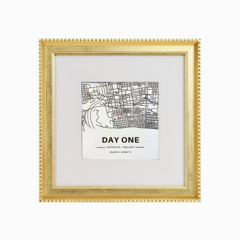 May include: A gold-colored picture frame with a white mat and a black and white map inside. The map shows a city with streets and a red dot marking a location. The text "DAY ONE" is printed below the map, along with the names "NICHOLAS + MELODY" and coordinates.