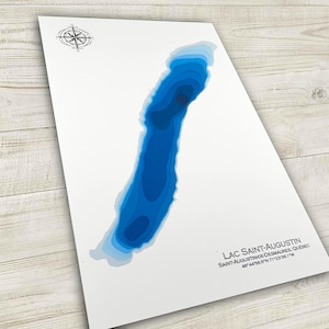 May include: A white print featuring a blue lake contour map with a compass rose in the upper left corner. The lake map is labeled "LAC SAINT-AUGUSTIN" and includes coordinates. The map is angled on a light wood surface.