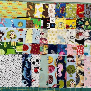 May include: A collection of fabric squares with various patterns, including musical notes, cartoon characters, animals, and food items. The fabrics are in a variety of colors, including green, blue, pink, and yellow. The squares are arranged on a green cutting mat.