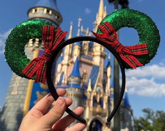 May include: Green and red Christmas-themed Minnie Mouse ears with a black headband. The ears are shaped like wreaths with red and green plaid bows.