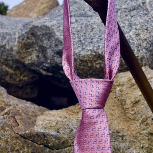May include: A pink silk tie with a blue pattern of small, repeated images of a seahorse. The tie is hanging from a wooden stick against a backdrop of large, gray rocks.