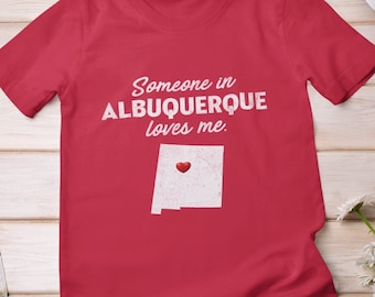 May include: Red t-shirt with a white graphic that says "Someone in Albuquerque loves me." and a white outline of the state of New Mexico with a red heart in the center.