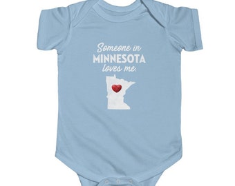 May include: Light blue baby onesie with white text that reads "Someone in Minnesota loves me."  A white outline of the state of Minnesota with a red heart in the center.