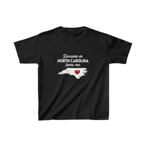 K&ouml;nnte beinhalten: Schwarzes T-Shirt mit wei&szlig;em Text, der "Jemand in North Carolina liebt mich" lautet. Der Text befindet sich &uuml;ber einem wei&szlig;en Umriss des Bundesstaates North Carolina mit einem roten Herzen in der Mitte.
