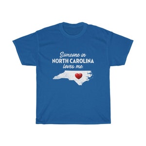 May include: Blue t-shirt with white text that reads "Someone in North Carolina loves me."  A red heart is inside the outline of the state of North Carolina.