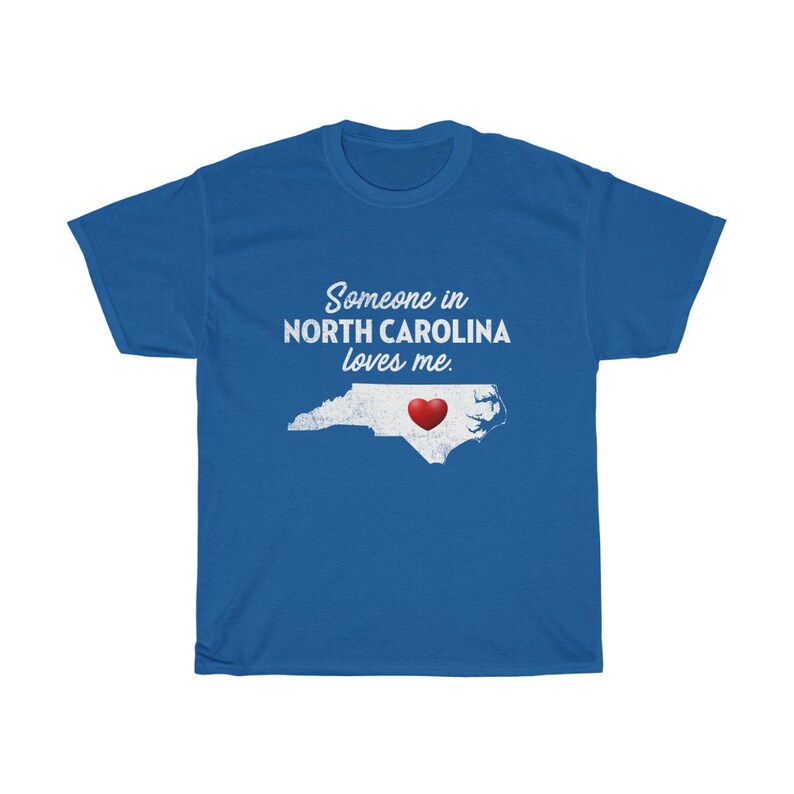May include: Blue t-shirt with white text that reads "Someone in North Carolina loves me."  A red heart is inside the outline of the state of North Carolina.