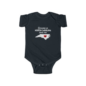 Peut inclure: Une grenouill&egrave;re b&eacute;b&eacute; bleu fonc&eacute; avec du texte blanc qui dit "Quelqu'un en Caroline du Nord m'aime." La grenouill&egrave;re pr&eacute;sente un contour blanc de l'&eacute;tat de Caroline du Nord avec un c&oelig;ur rouge au centre.