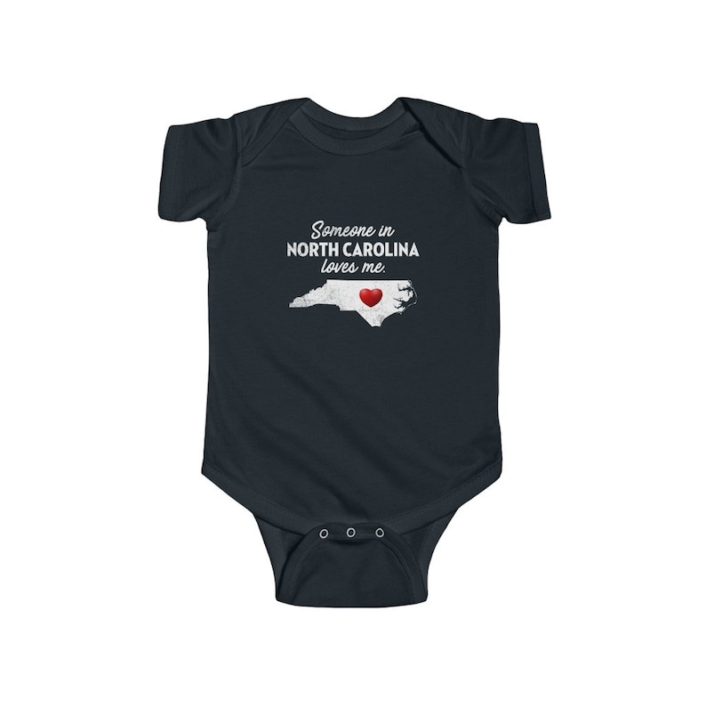Peut inclure: Une grenouill&egrave;re b&eacute;b&eacute; bleu fonc&eacute; avec du texte blanc qui dit "Quelqu'un en Caroline du Nord m'aime." La grenouill&egrave;re pr&eacute;sente un contour blanc de l'&eacute;tat de Caroline du Nord avec un c&oelig;ur rouge au centre.