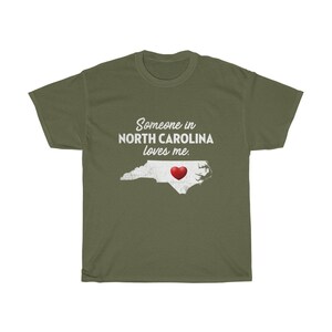 May include: Green t-shirt with white text that reads "Someone in North Carolina loves me."  The text is above an outline of the state of North Carolina with a red heart in the center.