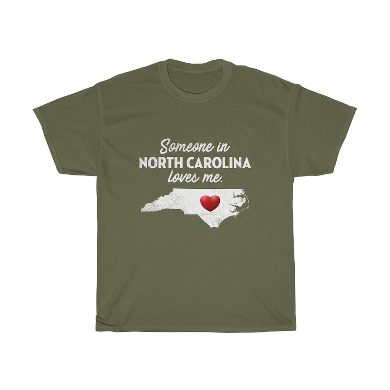 May include: Green t-shirt with white text that reads "Someone in North Carolina loves me."  The text is above an outline of the state of North Carolina with a red heart in the center.