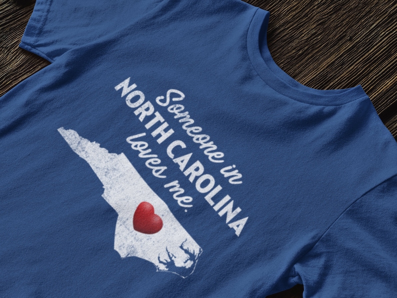 May include: A blue t-shirt with a white outline of the state of North Carolina and the text "Someone in North Carolina loves me." A red heart is inside the outline of the state.