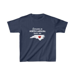 K&ouml;nnte beinhalten: Ein marineblaues T-Shirt mit einer wei&szlig;en Umrandung des Bundesstaates North Carolina und einem roten Herzen in der Mitte. Der Text "Someone in North Carolina loves me." ist auf das T-Shirt gedruckt.