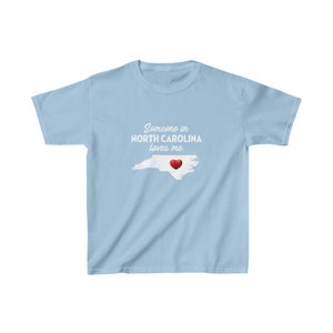 K&ouml;nnte beinhalten: Hellblaues T-Shirt mit einer wei&szlig;en Umrandung des Bundesstaates North Carolina und einem roten Herzen in der Mitte. Der Text "Someone in NORTH CAROLINA loves me." ist auf das T-Shirt gedruckt.