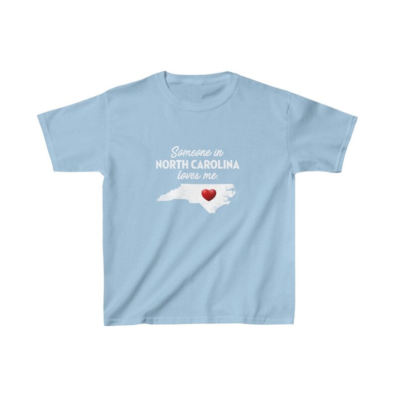 K&ouml;nnte beinhalten: Hellblaues T-Shirt mit einer wei&szlig;en Umrandung des Bundesstaates North Carolina und einem roten Herzen in der Mitte. Der Text "Someone in NORTH CAROLINA loves me." ist auf das T-Shirt gedruckt.