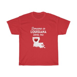 May include: Red t-shirt with white text that says "Someone in Louisiana loves me."  The text is over a white outline of the state of Louisiana with a red heart in the center.