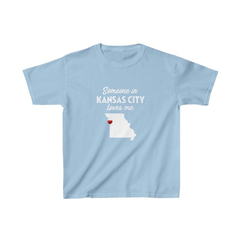 May include: Light blue t-shirt with white text that reads "Someone in Kansas City loves me." The text is centered on the shirt. A white outline of the state of Missouri is below the text with a red heart over Kansas City.