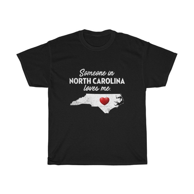 May include: Black t-shirt with white text that reads "Someone in North Carolina loves me."  The text is in a vintage style font.  A white outline of the state of North Carolina with a red heart in the center is below the text.