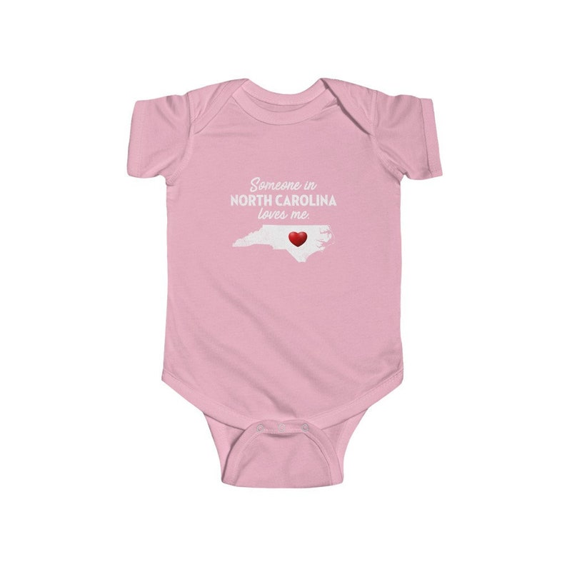 Peut inclure: Combinaison rose pour b&eacute;b&eacute; avec un texte blanc qui dit "Quelqu'un en Caroline du Nord m'aime". La combinaison pr&eacute;sente un contour blanc de l'&eacute;tat de Caroline du Nord avec un c&oelig;ur rouge au centre.