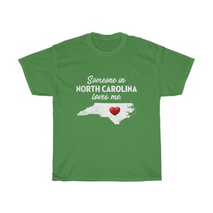 May include: Green t-shirt with white text that reads "Someone in North Carolina loves me."  A white outline of the state of North Carolina with a red heart in the center.