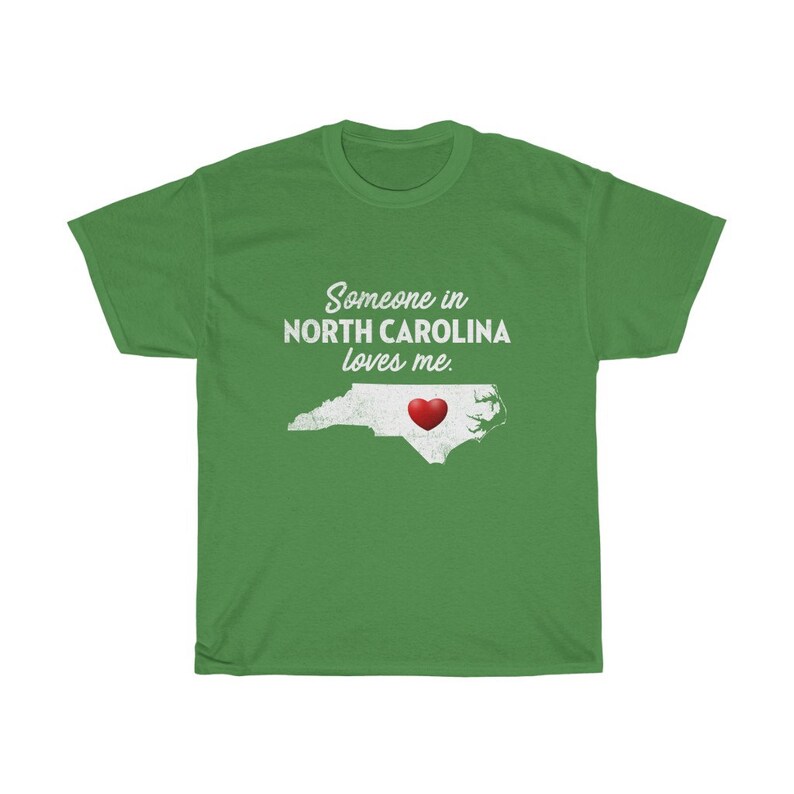 May include: Green t-shirt with white text that reads "Someone in North Carolina loves me."  A white outline of the state of North Carolina with a red heart in the center.