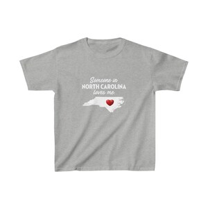 K&ouml;nnte beinhalten: Graues T-Shirt mit einer wei&szlig;en Umrandung von North Carolina und einem roten Herzen in der Mitte. Der Text "Jemand in North Carolina liebt mich" ist auf das T-Shirt gedruckt.