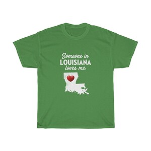 May include: Green t-shirt with white text that reads "Someone in LOUISIANA loves me."  A white outline of the state of Louisiana with a red heart in the center is below the text.