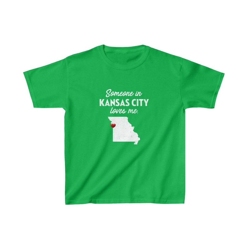 May include: Green t-shirt with white text that reads "Someone in Kansas City loves me."  The text is centered on the shirt.  A white outline of the state of Missouri with a red heart in the location of Kansas City is below the text.