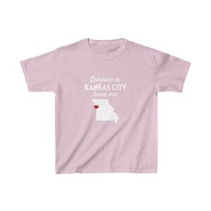 May include: Pink t-shirt with a white outline of the state of Missouri and the text "Someone in Kansas City loves me." A red heart is placed over Kansas City.