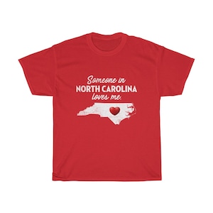 May include: Red t-shirt with white text that reads "Someone in North Carolina loves me."  A white outline of the state of North Carolina with a red heart in the center.