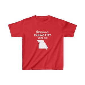 May include: Red youth t-shirt with white text that reads "Someone in Kansas City loves me."  The shirt features a white outline of the state of Missouri with a red heart in the location of Kansas City.