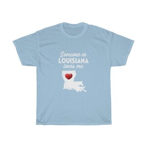 May include: Light blue t-shirt with the text "Someone in LOUISIANA loves me." and a white outline of the state of Louisiana with a red heart in the center.