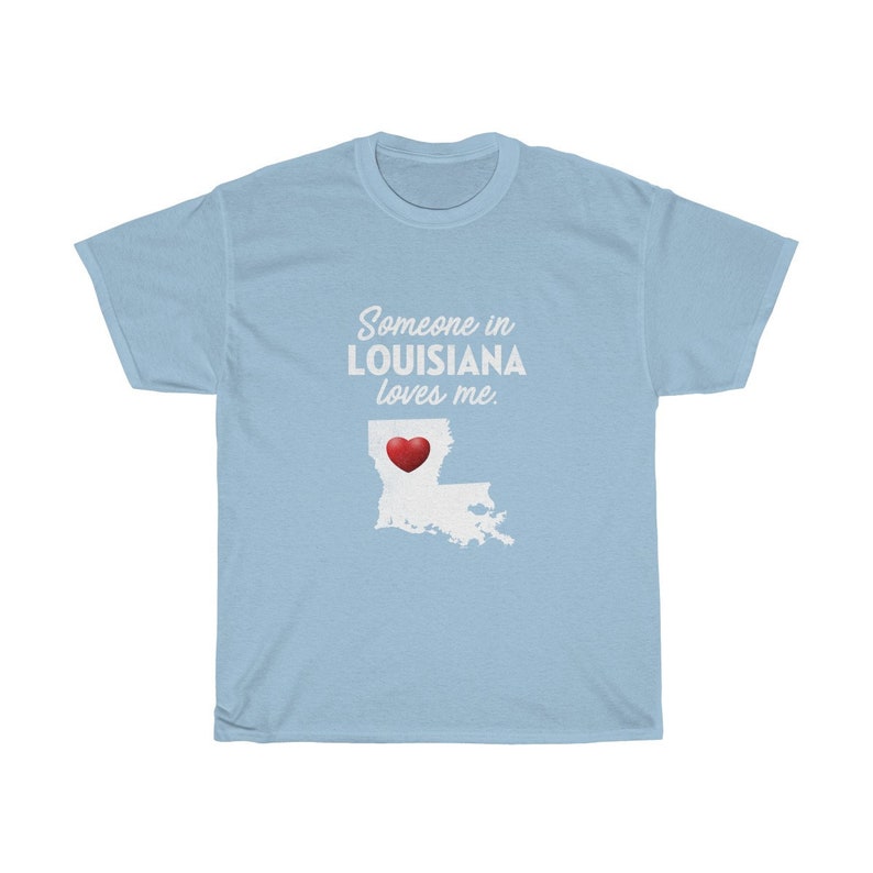 May include: Light blue t-shirt with the text "Someone in LOUISIANA loves me." and a white outline of the state of Louisiana with a red heart in the center.