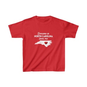 K&ouml;nnte beinhalten: Rotes T-Shirt mit einer wei&szlig;en Umrandung von North Carolina und einem roten Herzen in der Mitte. Der Text "Jemand in North Carolina liebt mich." ist auf das T-Shirt gedruckt.