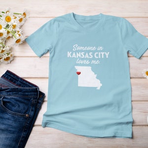 Puede incluir: Camiseta azul claro con el texto "Someone in KANSAS CITY loves me." y un contorno blanco del estado de Misuri con un coraz&oacute;n rojo sobre Kansas City.
