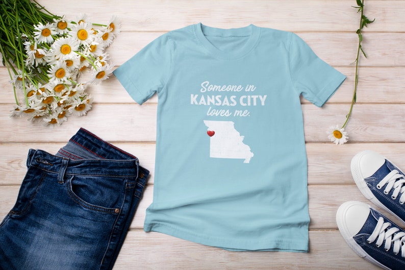 Puede incluir: Camiseta azul claro con el texto "Someone in KANSAS CITY loves me." y un contorno blanco del estado de Misuri con un coraz&oacute;n rojo sobre Kansas City.