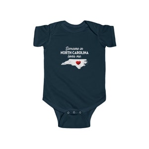 Peut inclure: Combinaison b&eacute;b&eacute; bleu marine avec un contour blanc de l'&eacute;tat de Caroline du Nord et un c&oelig;ur rouge au centre. Le texte "Someone in NORTH CAROLINA loves me." est imprim&eacute; sur la combinaison.