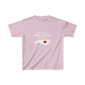 K&ouml;nnte beinhalten: Rosafarbenes T-Shirt mit einer wei&szlig;en Umrandung des Bundesstaates North Carolina und einem roten Herzen in der Mitte. Der Text "Jemand in North Carolina liebt mich." ist auf das T-Shirt gedruckt.
