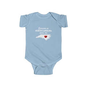 Peut inclure: Combinaison b&eacute;b&eacute; bleu clair avec un texte blanc qui dit "Quelqu'un en Caroline du Nord m'aime." Le texte est au-dessus d'un contour de l'&eacute;tat de Caroline du Nord avec un c&oelig;ur rouge au centre.