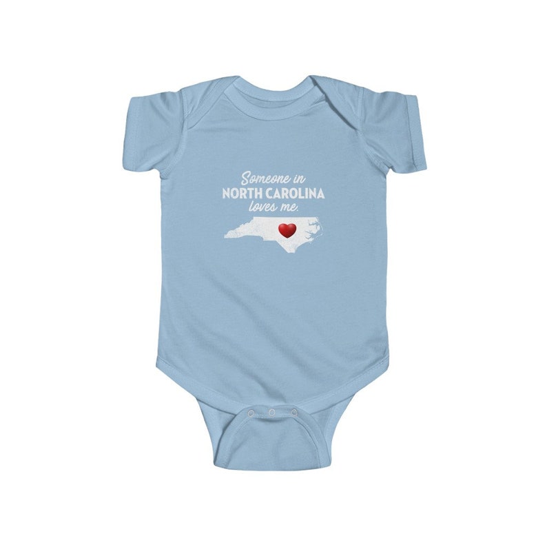 Peut inclure: Combinaison b&eacute;b&eacute; bleu clair avec un texte blanc qui dit "Quelqu'un en Caroline du Nord m'aime." Le texte est au-dessus d'un contour de l'&eacute;tat de Caroline du Nord avec un c&oelig;ur rouge au centre.