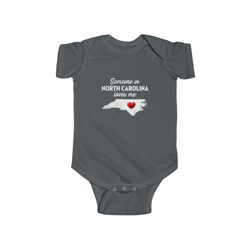 Peut inclure: Une grenouill&egrave;re b&eacute;b&eacute; gris fonc&eacute; avec un texte blanc qui dit "Quelqu'un en CAROLINE DU NORD m'aime." La grenouill&egrave;re pr&eacute;sente un contour blanc de l'&eacute;tat de Caroline du Nord avec un c&oelig;ur rouge au centre.