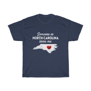 May include: Navy blue t-shirt with a white outline of the state of North Carolina and a red heart in the center. The text "Someone in North Carolina loves me." is printed in white on the shirt.