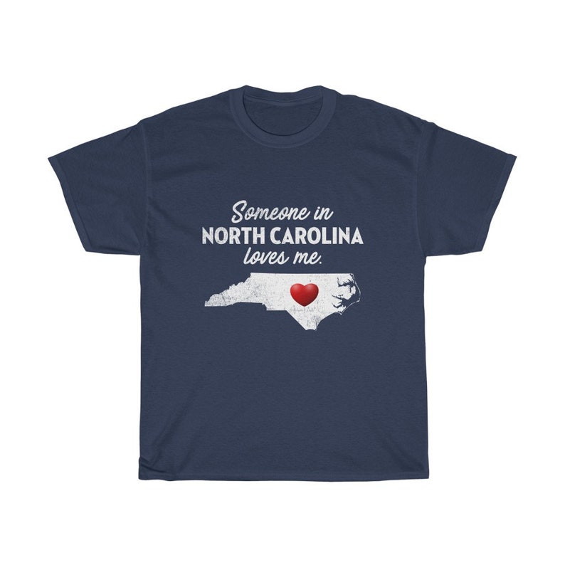 May include: Navy blue t-shirt with a white outline of the state of North Carolina and a red heart in the center. The text "Someone in North Carolina loves me." is printed in white on the shirt.