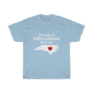 May include: Light blue t-shirt with a white outline of the state of North Carolina and a red heart in the center. The text "Someone in North Carolina loves me." is printed in white on the shirt.