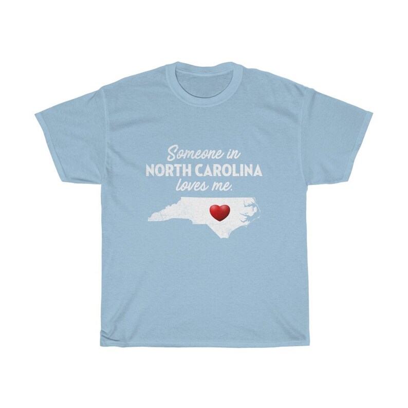 May include: Light blue t-shirt with a white outline of the state of North Carolina and a red heart in the center. The text "Someone in North Carolina loves me." is printed in white on the shirt.