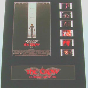 May include: A black framed movie poster for the film "The Crow" featuring actor Brandon Lee. The poster has a red crow logo and the text "The Crow" in red. The poster also includes the text "U.S. Release - May 12, 1994". The poster is accompanied by a set of film cell strips.