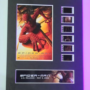 May include: A framed movie poster and film cell display featuring Spider-Man. The poster is red, blue, and black with the title "Spider-Man" and the release date "May 3, 2002". The film cells are in black and white and show scenes from the movie.