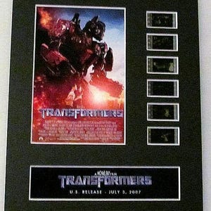 May include: A framed movie poster for the 2007 film Transformers, featuring a black robot with red accents. The poster is surrounded by a black frame with a black mat. The title of the film is written in white letters on the poster. The poster also includes the text "A Michael Bay Film" and "U.S. Release - July 3, 2007".