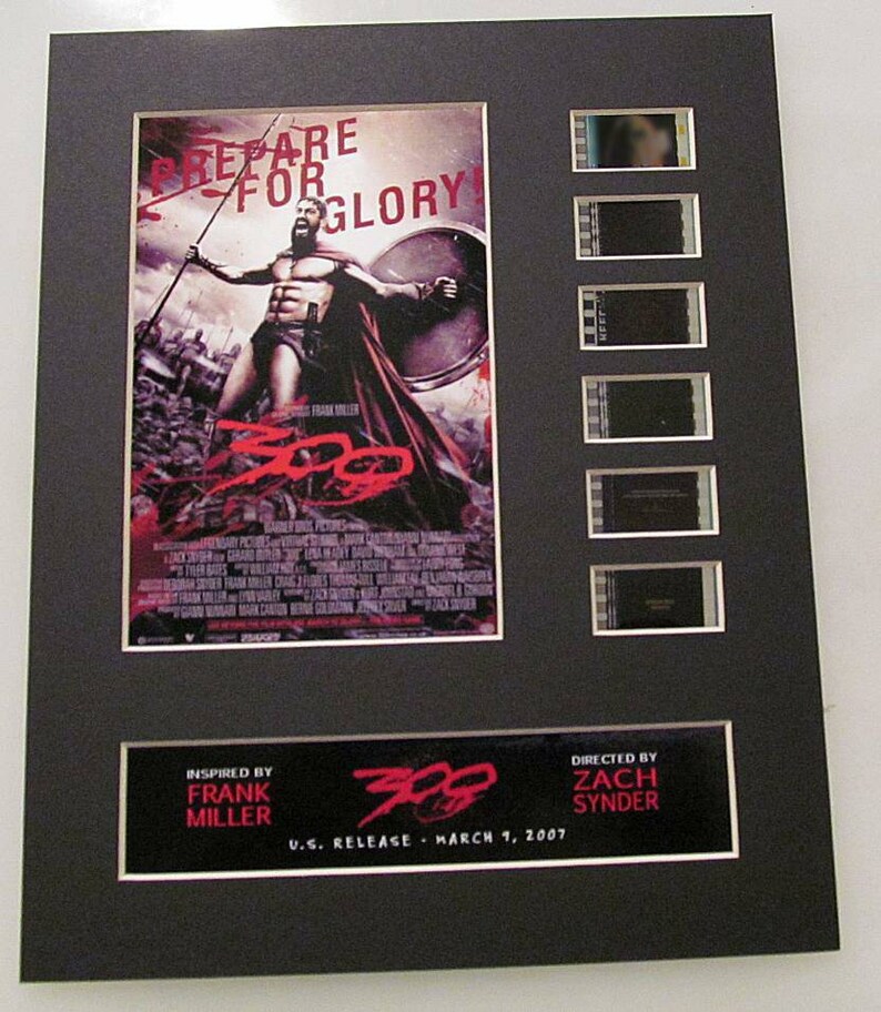 May include: A framed movie poster for the film "300" with a red and black color scheme. The poster features a scene from the movie with a warrior holding a spear. The poster also includes the text "Prepare for Glory!" and "Inspired by Frank Miller". The poster is accompanied by five film strips.